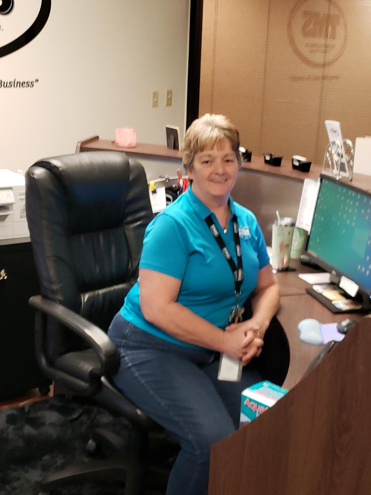 a woman smiling while she sits at a desk with a computer
